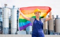 Young female engineer in helmet waving rainbow LGBT flag while standing in front of big tanks at chemical plant