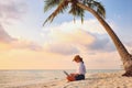 Young female freelancer wearing straw hat working on laptop while sitting on tropical beach at sunset