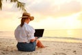 Young female freelancer wearing straw hat working on laptop while sitting on tropical beach at sunset Royalty Free Stock Photo