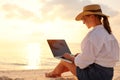 Young female freelancer wearing straw hat working on laptop while sitting on tropical beach at sunset Royalty Free Stock Photo