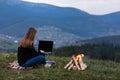 Young female freelancer working on laptop in the mountains in the evening. Tourist girl sitting near campfire and having fun. Copy