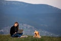Young female freelancer working on laptop in the mountains in the evening. Tourist girl sitting near campfire and having fun. Copy
