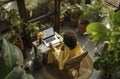 Young female gardener using laptop, communicates on internet with customer in home garden greenhouse, reusable coffee tea mug on