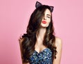 Young female gives kiss shows red lips with make up wears retro style cat ears modern pink background pinup girl