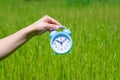 Young female hand holds sunlit alarm clock on green blurred nackground with bokeh effect