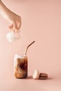 Young female hand pouring milk in glass of iced coffee in tall glass with golden straw. Coffee and macaroons chocolate