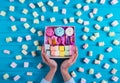 Young female hands holding box full of colorful macaroons and marshmallow with a lot of square different marshmallow on blue