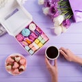 Young female hands holding cup of coffee near colorful marshmallow and macaroon set in box, bouquet of purple and white tulips and Royalty Free Stock Photo