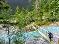 A young female hiker carefully crossing a small skinny wooden bridge over a narrow electric blue rushing river