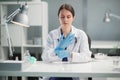 A young female internist is sitting at a desk in her spacious, bright office, wearing gloves and a medical gown