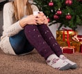 Young girl sitting on floor and holding cup of coffee in hands on legs with warmers and Christmas tree in background Royalty Free Stock Photo