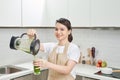 Young female in linen apron pouring green smoothie drink from blender to bottle surrounded with vegetables and greens