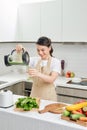 Young female in linen apron pouring green smoothie drink from blender to bottle surrounded with vegetables and greens
