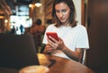 Young female manager holding smartphone using laptop in cafe, hipster girl freelancer writing on mobile phone, businesswoman work Royalty Free Stock Photo