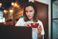 Young female manager using laptop in cafe, hipster girl freelancer looking on monitor and drink hot coffee, businesswoman working Royalty Free Stock Photo