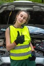 Young female mechanic in green uniform standing near a broken-down car and waiting help