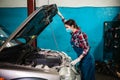 A young female mechanic in a uniform and medical mask opens the hood of a car. Side view. Indoors garage