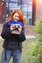 Young female photographer looking at her camera with red hair is taking pictures in the city on a walk. City landscape background Royalty Free Stock Photo