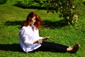 Young female poetess with long red hair thinking about read exciting story. Sitting in grass with book. Woman enjoying