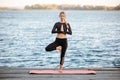 Young female practicing yoga with a mat on a wooden pier near the calm blue sea Royalty Free Stock Photo