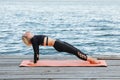 Young female practicing yoga with a mat on a wooden pier near the calm blue sea Royalty Free Stock Photo