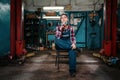 A young female pretty mechanic in a work uniform is sitting on a chair, with a medical mask on her chin. Indoor. Garage