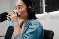 Young female radio host with headphones drinking coffee in studio