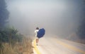 Young female in a raincoat and umbrella on the road in the fog. Travel of women in the raincoat hitchhiking in the rain Royalty Free Stock Photo