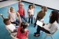 Young female school teacher reading a book to infant school children, sitting on chairs in a circle in the classroom raising hands