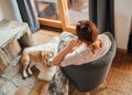 Young female sitting in comfortable chair in country house living room and dreaming and her beagle dog lying near feet on the