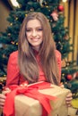 Young female is sitting in front of the christmas tree and have gift box with red ribbon in her hands