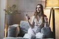 Young female sitting on sofa with smartphone in hand, wearing earphones and listening to music. Girl with mobile phone sending Royalty Free Stock Photo