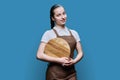 Young female student waitress in an apron with wooden empty tray on blue background