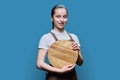 Young female student waitress in an apron with wooden empty tray on blue background