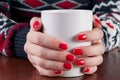 Young girl in sweater holding white coffee cup in hand with red nails on wooden desk Royalty Free Stock Photo