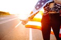 Young female tourist with american flag hitchhiking along a highway. Close up woman`s hand showind thumbs up. Face is Royalty Free Stock Photo