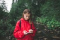 Young female traveler in a red raincoat using smartphone in a mountain forest. Beautiful caucasian hiker girl in red jacket typing