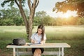 Young female university student writes a note and assignment with a laptop beside outside the campus with blur green tree backgrou