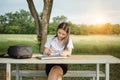 Young female university student writes a note and assignment with a laptop beside outside the campus with blur green tree backgrou