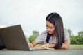 Young female university student writes a note and assignment with a laptop beside outside the campus with blur green tree backgrou