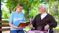 Young female volunteer reading book for disabled pensioner in wheelchair, care Royalty Free Stock Photo