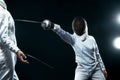 Young fencer athlete wearing mask and white fencing costume. holding the sword on black background with lights.