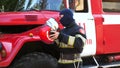 Young fireguard taking off helmet and balaclava stands near fire engine. Male firefighter removing equipment from