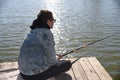 Young fisherman in denim jacket sitting on wooden pier, fishing in the lake. he holding the rod and looking at float under bright Royalty Free Stock Photo