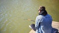 Young fisherman in denim jacket sitting on wooden pier, fishing in the lake. he holding the rod and looking at float. Royalty Free Stock Photo