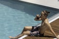 Young fit man sitting on the edge of the swimming pool relaxing while drinking a coke beverage and enjoying the weather