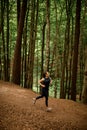 Young fit woman in black sportswear jogging downhill on forest trail, tall leafy trees on background