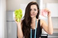 Young fit woman with centimeter round her neck holding fresh salad in one hand and slice of ham in another looks hesitant ,