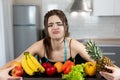 Young fit woman with centimeter round neck wearing black sports top standing in the kitchen full of fruits looks upset dietology