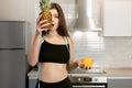 Young fit woman with centimeter round neck wearing black top and leggings holds pineapple near her face standing in kitchen